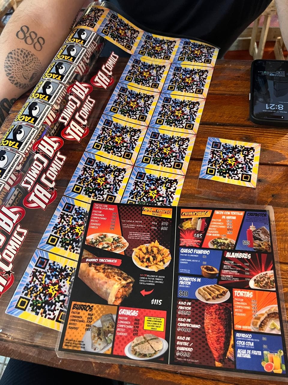 Tattooed arm beside a colorful Mexican menu and a sheet of vibrant QR code stickers.