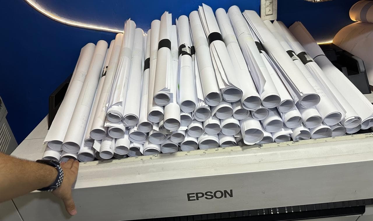 Rolled white blueprints stacked high on an Epson wide-format printer in a blue room.